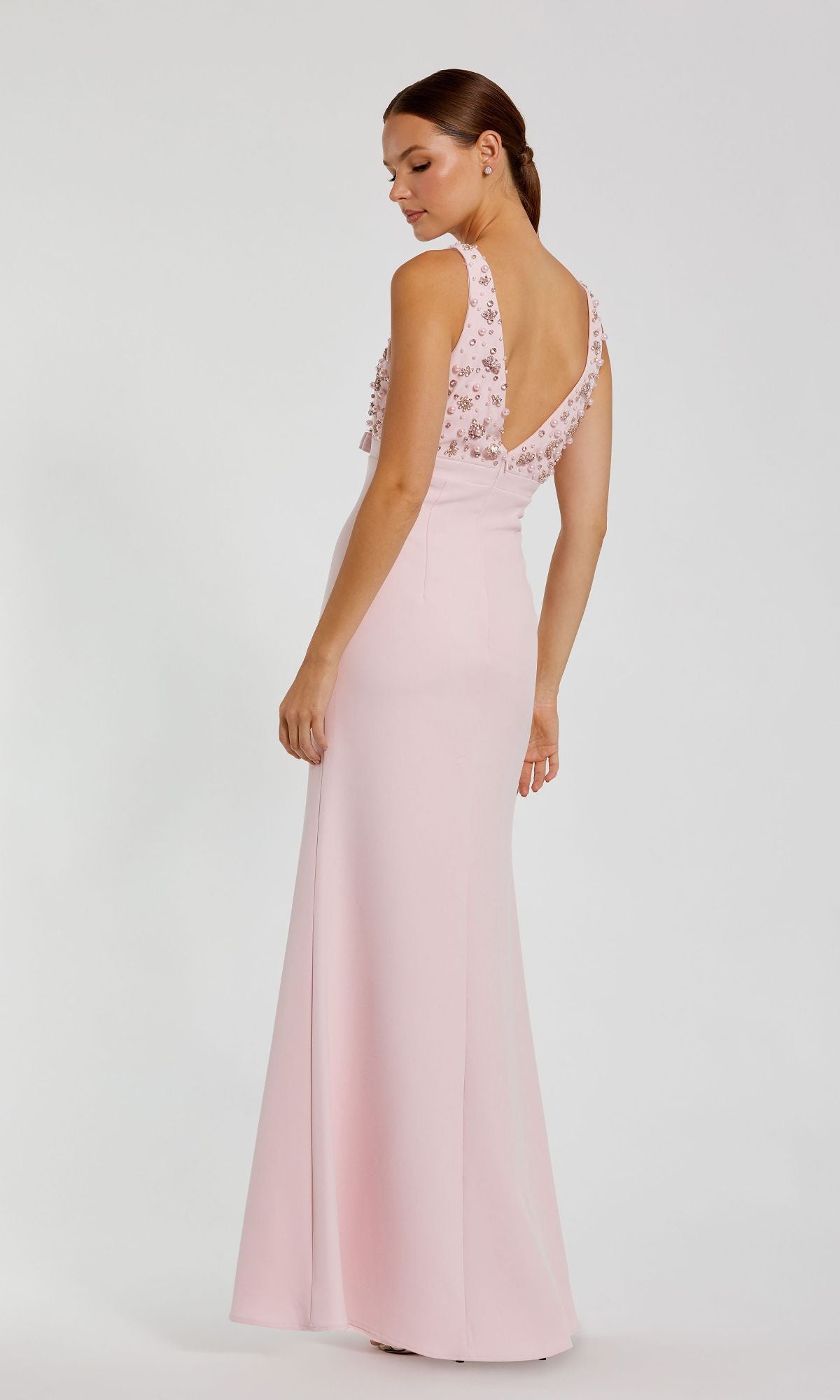 A woman poses against a plain light background in the Mac Duggal 2278 Beaded-Bodice Long Prom Dress, a light pink gown with floral embellishments and a deep V-back.